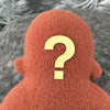 A small orange fleece figure with a yellow question mark where its face should be with a blurred white textured background,