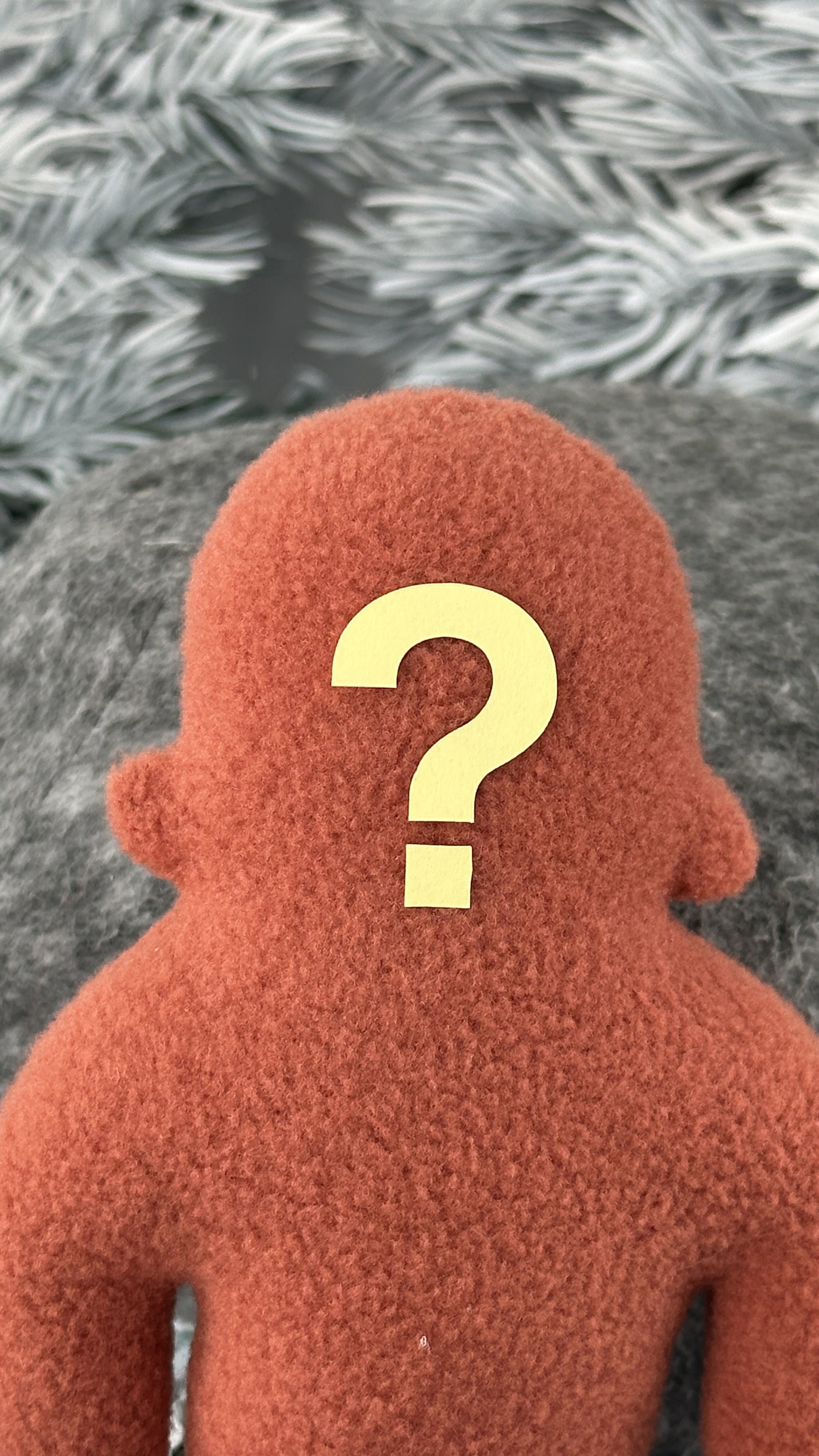 A small orange fleece figure with a yellow question mark where its face should be with a blurred white textured background,