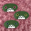 Three plush, green, oval-shaped patches with jagged mouths and colorful eyes are set against a textured, pink background resembling fur.