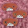 Three plush, orange, oval-shaped patches with jagged mouths and colorful eyes are set against a textured, pink background resembling fur.