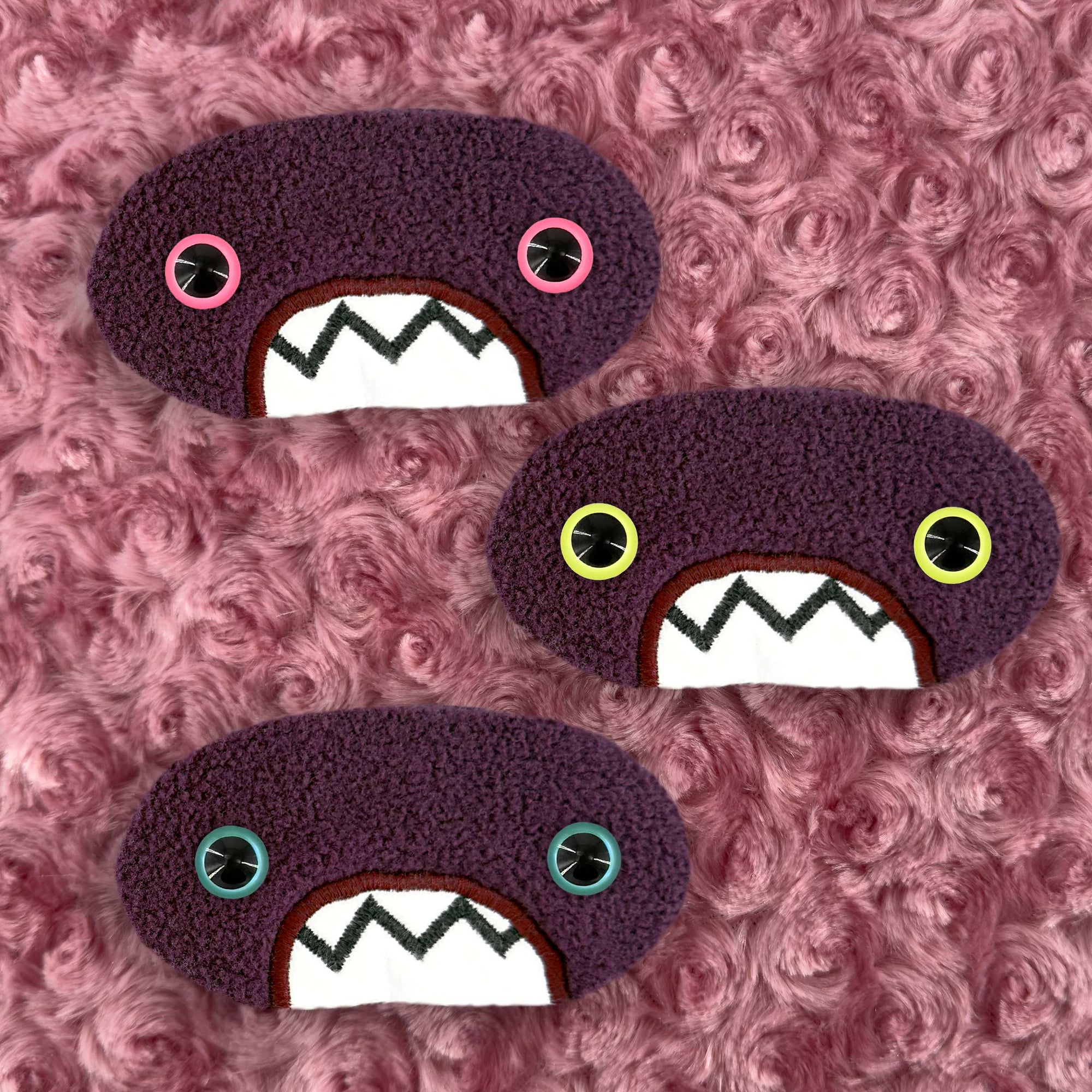 Three plush, purple, oval-shaped patches with jagged mouths and colorful eyes are set against a textured, pink background resembling fur.