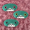 Three plush, teal, oval-shaped patches with jagged mouths and colorful eyes are set against a textured, pink background resembling fur.