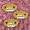 Three plush, mustard colored oval-shaped patches with jagged mouths and colorful eyes are set against a textured, pink background resembling fur.