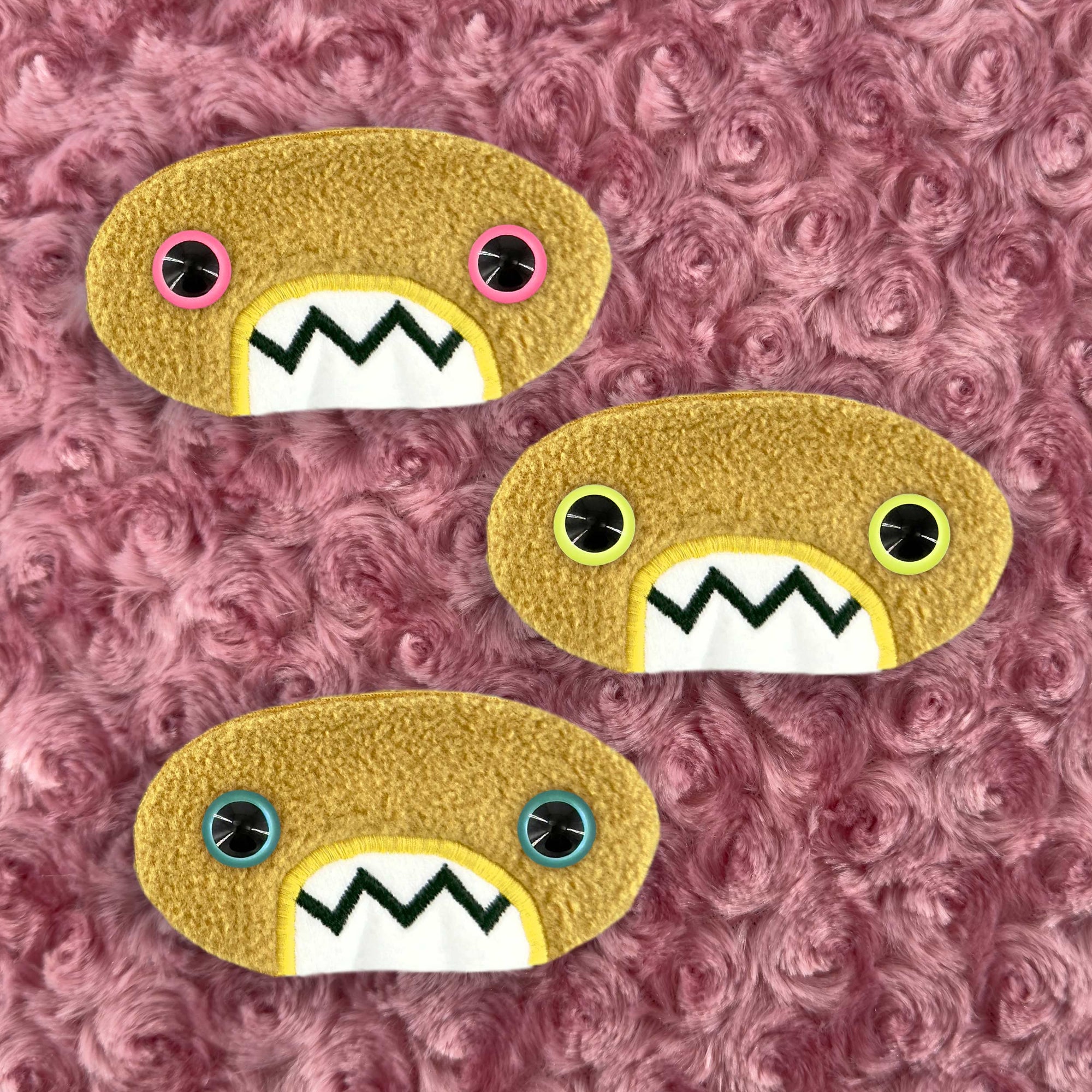 Three plush, mustard colored oval-shaped patches with jagged mouths and colorful eyes are set against a textured, pink background resembling fur.