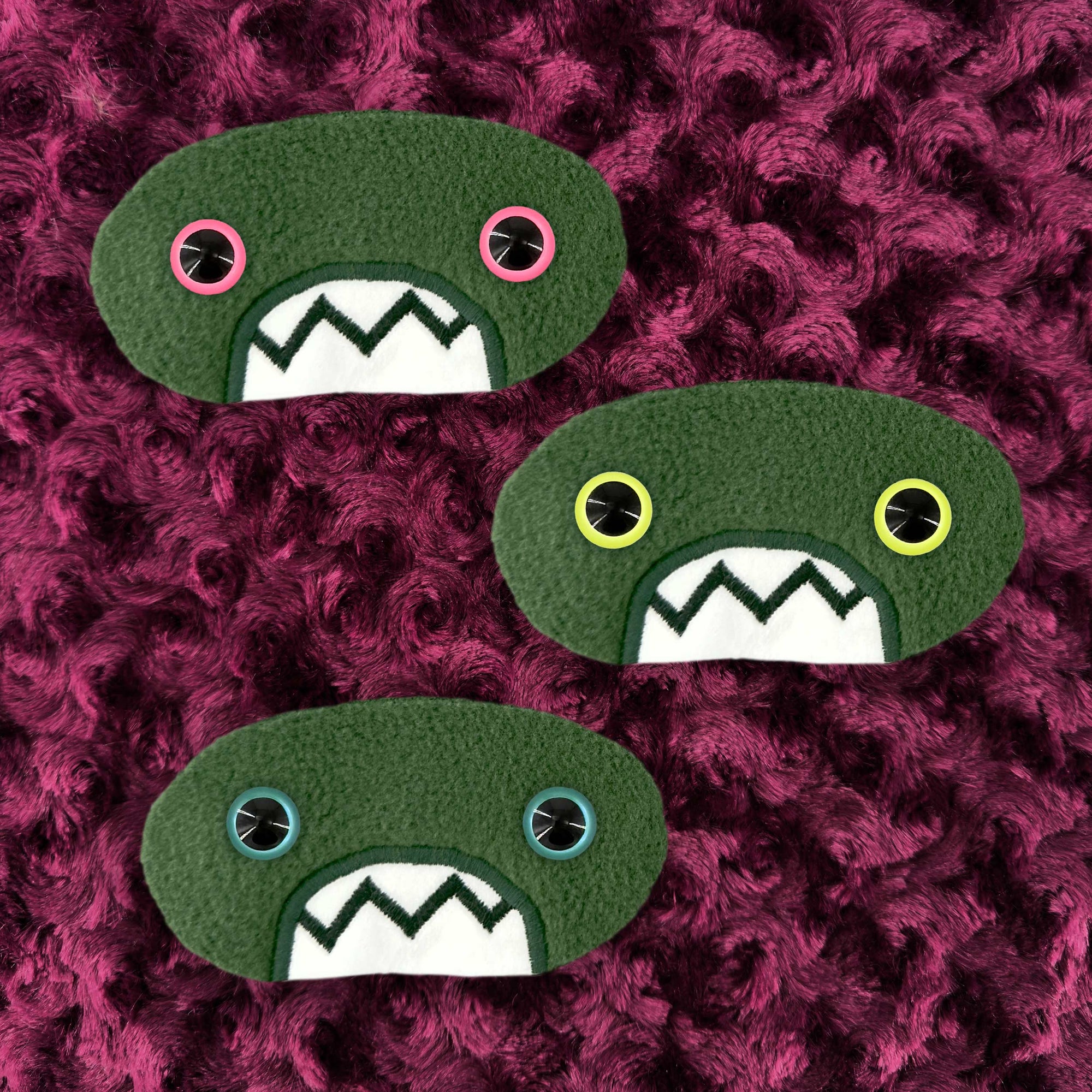 Three plush, green, oval-shaped patches with jagged mouths and colorful eyes are set against a textured, burgundy background resembling fur.