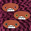 Three plush, orange, oval-shaped patches with jagged mouths and colorful eyes are set against a textured, burgundy background resembling fur.