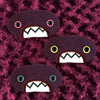 Three plush, purple, oval-shaped patches with jagged mouths and colorful eyes are set against a textured, burgundy background resembling fur.