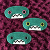 Three plush, teal, oval-shaped patches with jagged mouths and colorful eyes are set against a textured, burgundy background resembling fur.