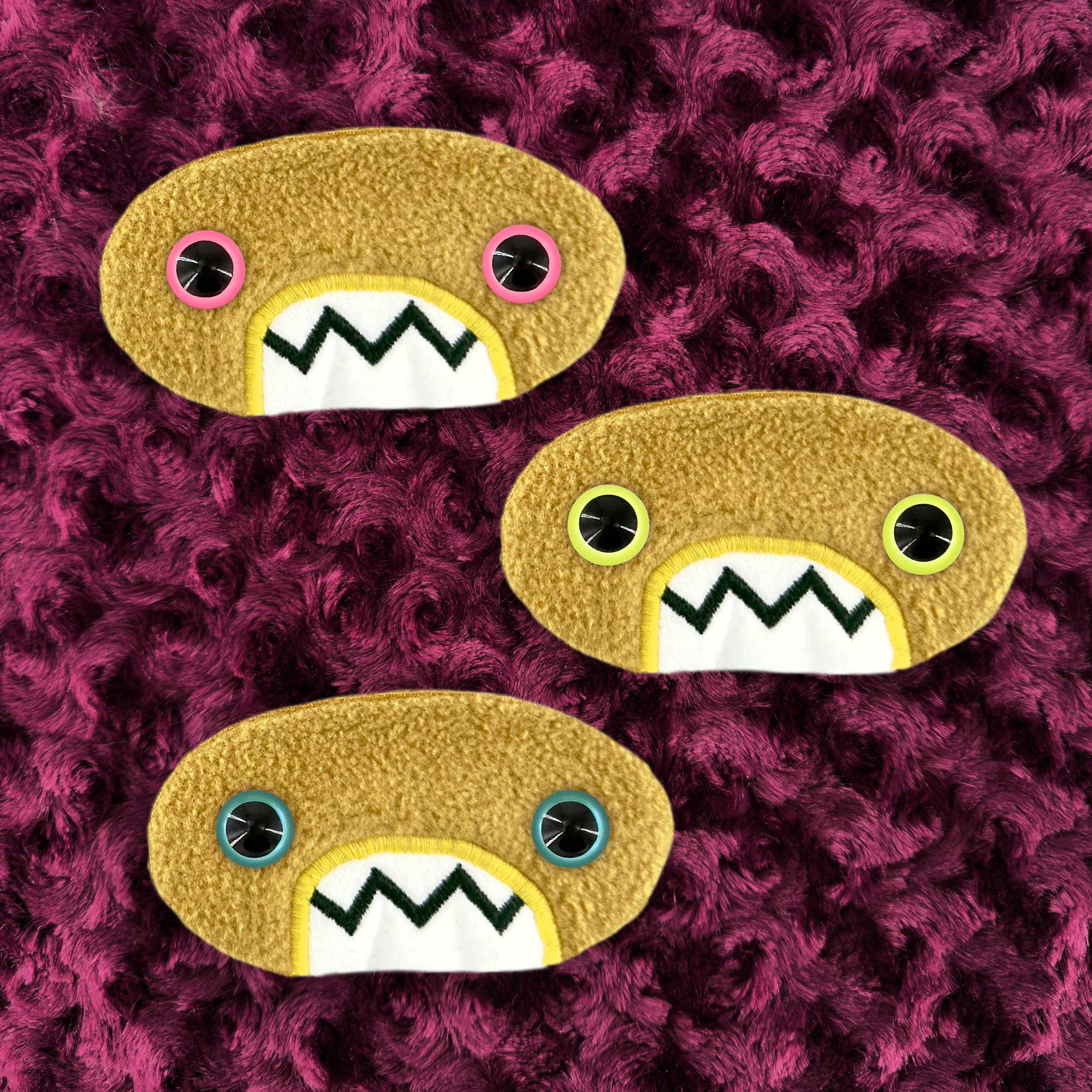 Three plush, mustard colored oval-shaped patches with jagged mouths and colorful eyes are set against a textured, burgundy background resembling fur.