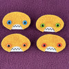 Four oval shaped orange faces with different eye colors and pointed teeth sit atop a purple fleece background.