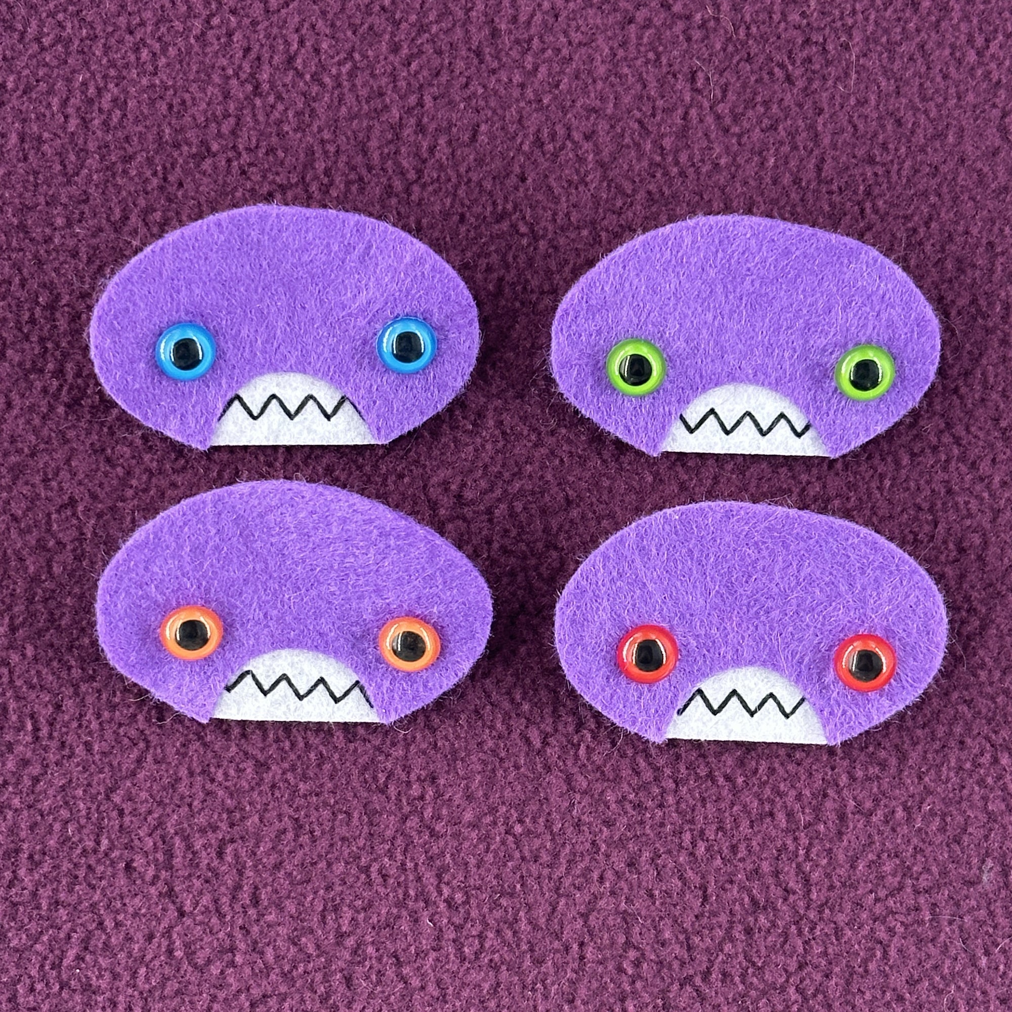 Four oval shaped purple faces with different eye colors and pointed teeth sit atop a purple fleece background.