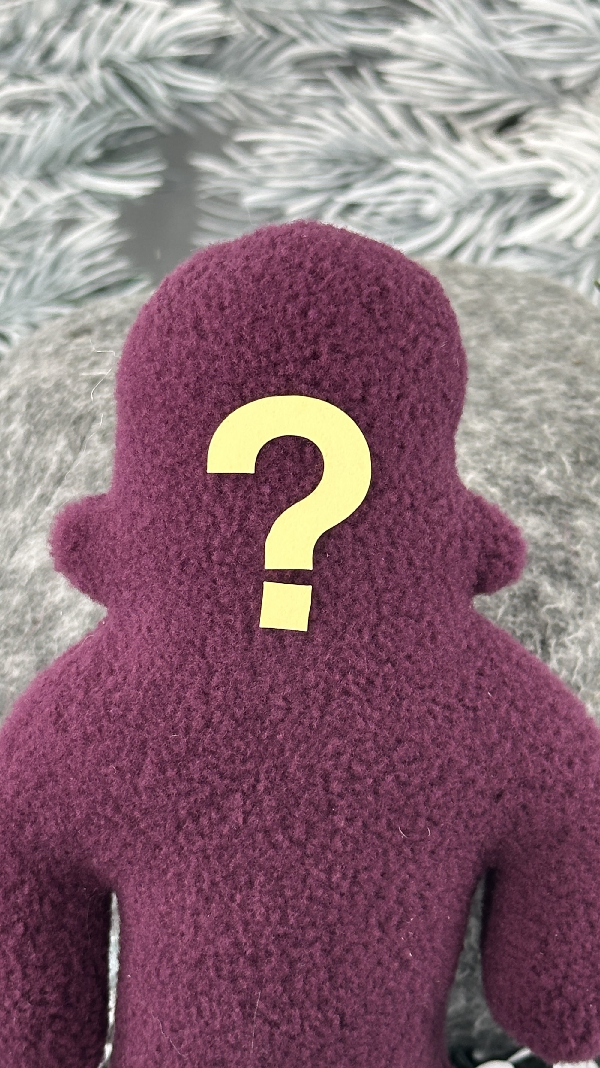 A small purple fleece figure with a question mark face stands in front of white foliage.