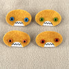 Four oval shaped orange faces with different eye colors and pointed teeth sit atop a sand colored fleece background.