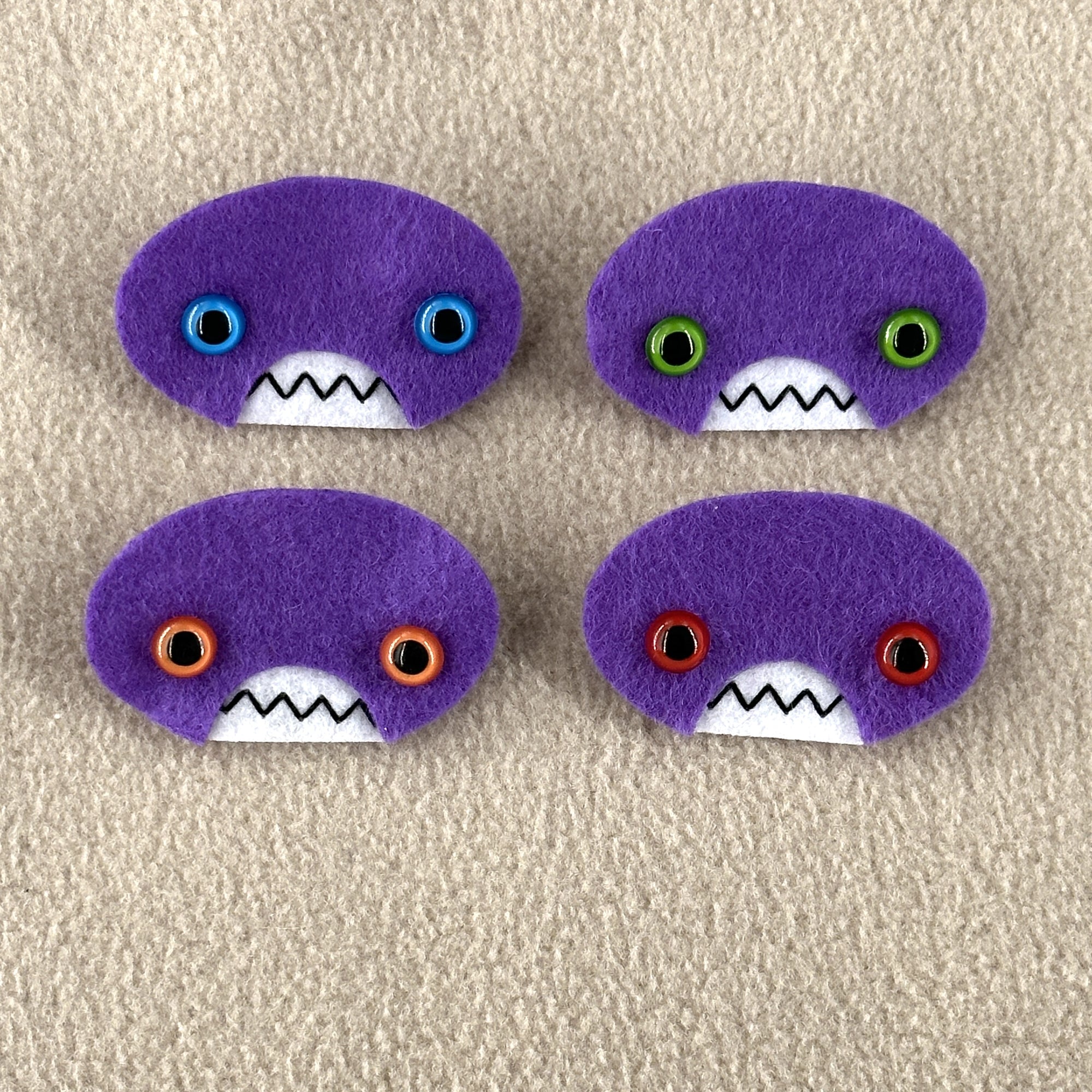 Four oval shaped purple faces with different eye colors and pointed teeth sit atop a sand colored fleece background.