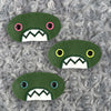 Three plush, green, oval-shaped patches with jagged mouths and colorful eyes are set against a textured, silver background resembling fur.