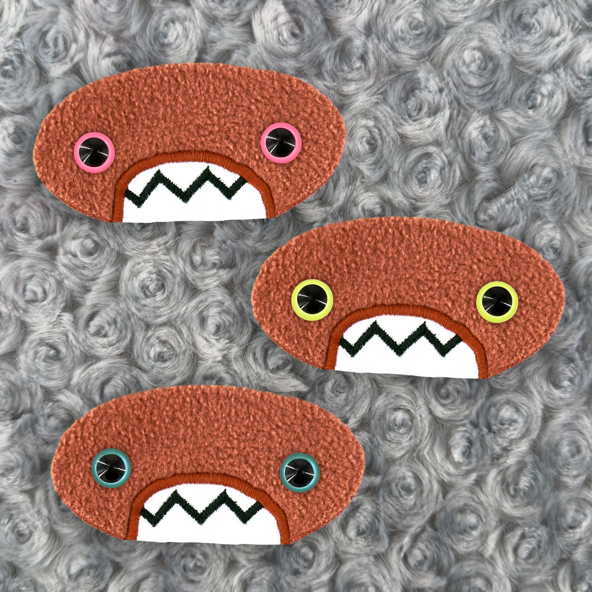 Three plush, orange, oval-shaped patches with jagged mouths and colorful eyes are set against a textured, silver background resembling fur.