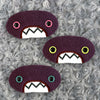 Three plush, purple, oval-shaped patches with jagged mouths and colorful eyes are set against a textured, silver background resembling fur.