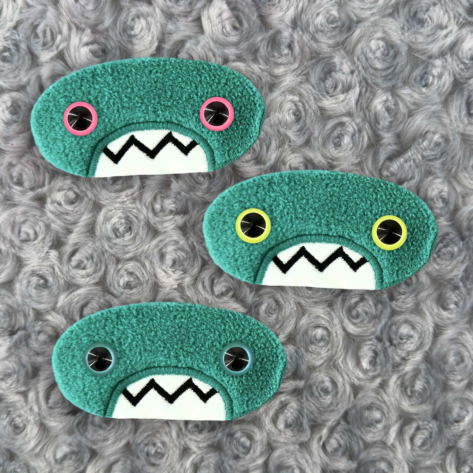 Three plush, teal, oval-shaped patches with jagged mouths and colorful eyes are set against a textured, silver background resembling fur.