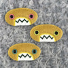 Three plush, mustard colored oval-shaped patches with jagged mouths and colorful eyes are set against a textured, silver background resembling fur.