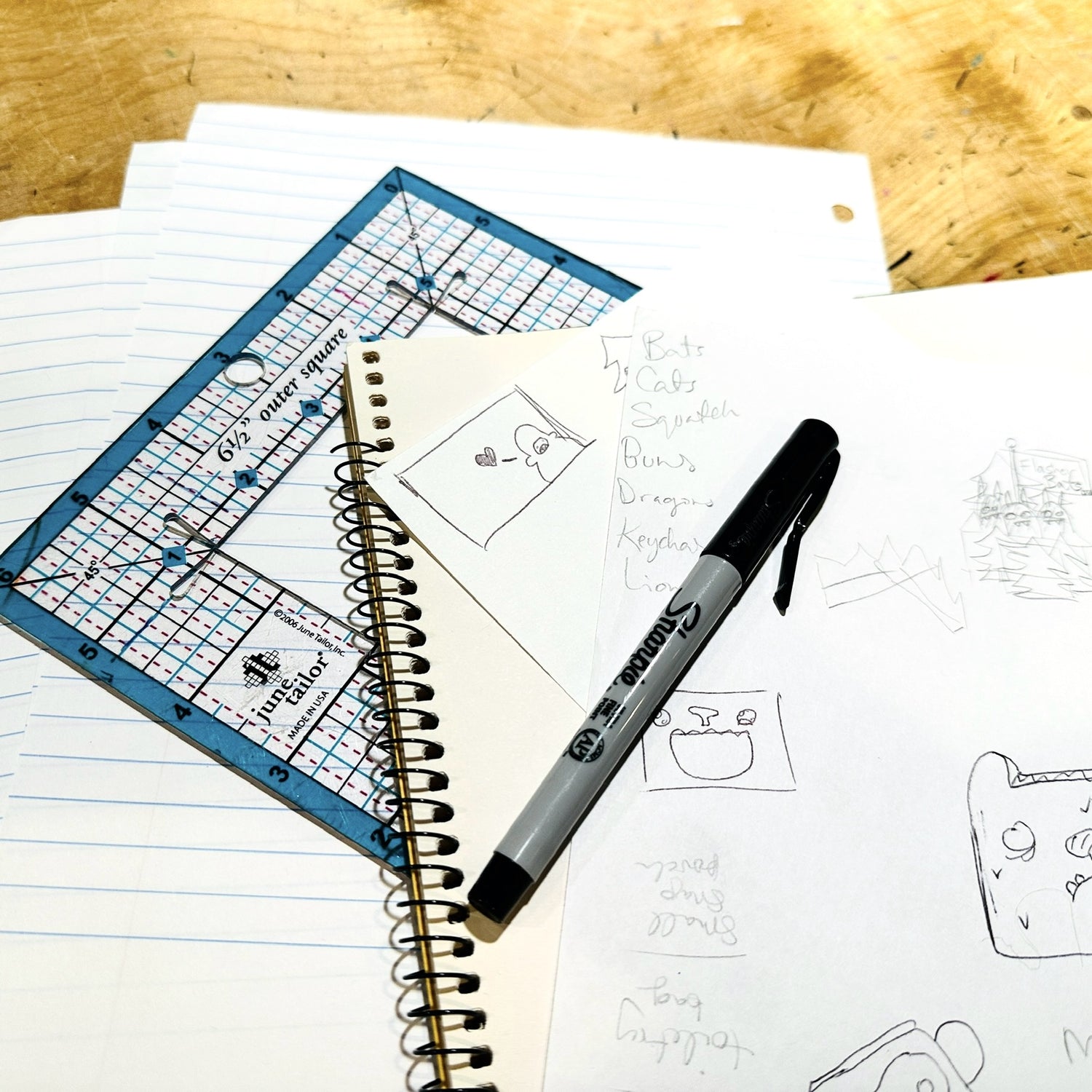 Scattered papers with sketches are layered with a market and square ruler utop a wooden table.