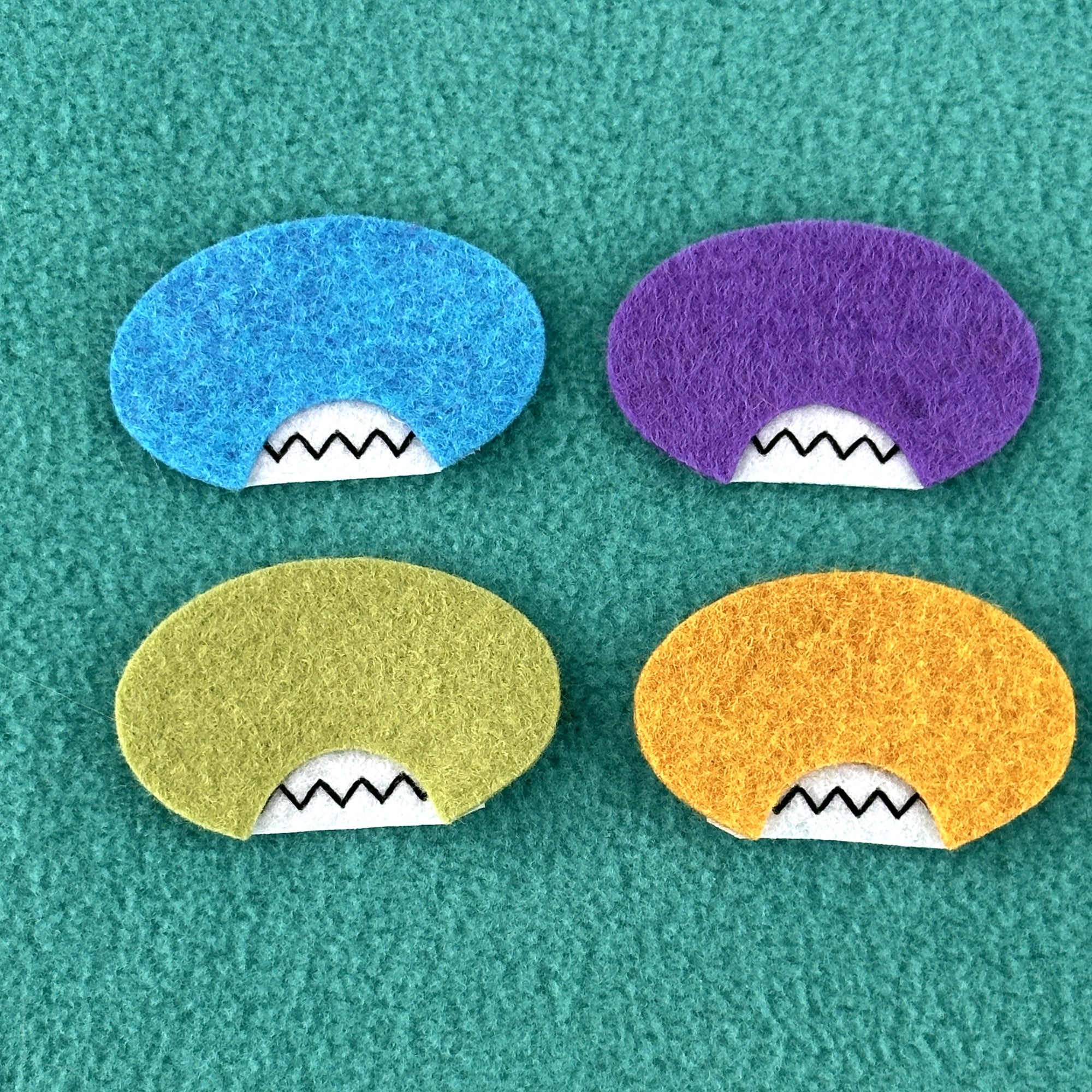 A group of four different colored oval felt faces with mouths and no eyes sit on at teal fleece background.