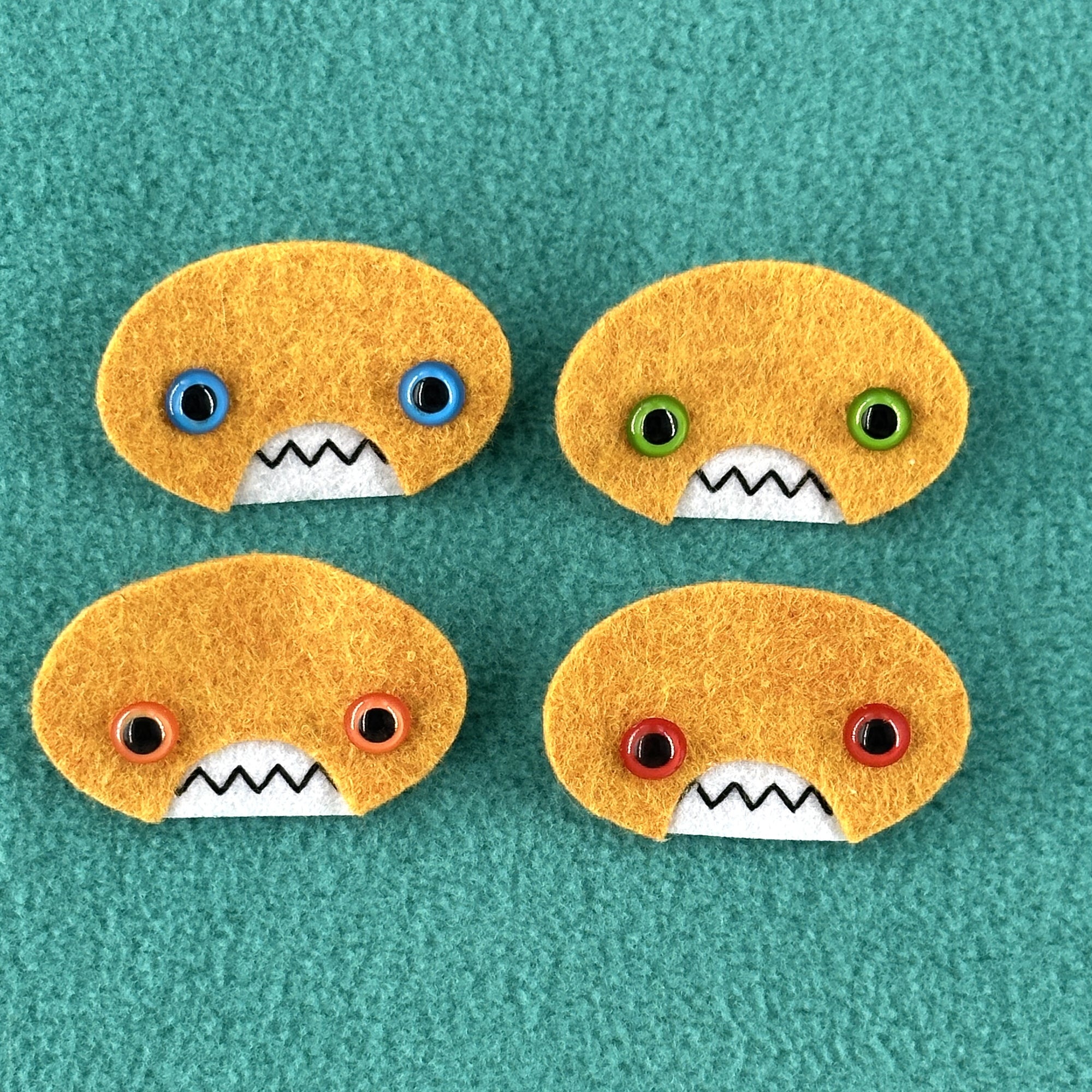 Four oval shaped orange faces with different eye colors and pointed teeth sit atop a teal fleece background.