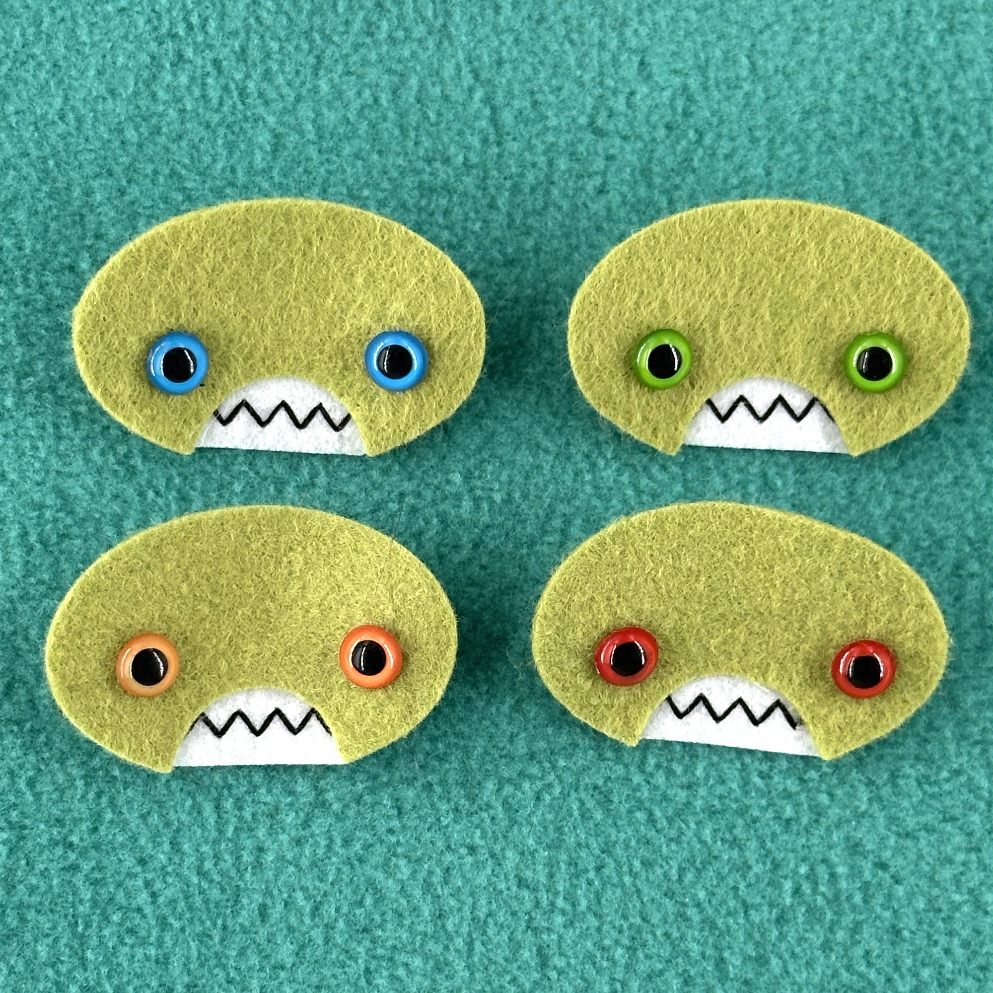 Four oval shaped green faces with different eye colors and pointed teeth sit atop a teal fleece background.