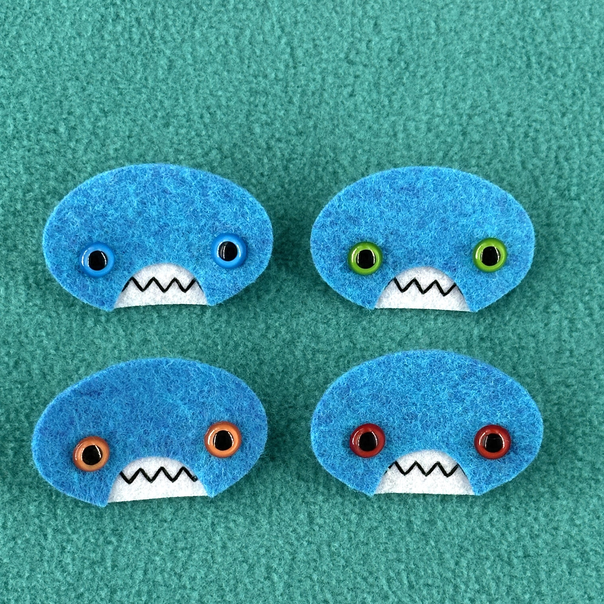 Four oval shaped teal faces with different eye colors and pointed teeth sit atop a teal fleece background.