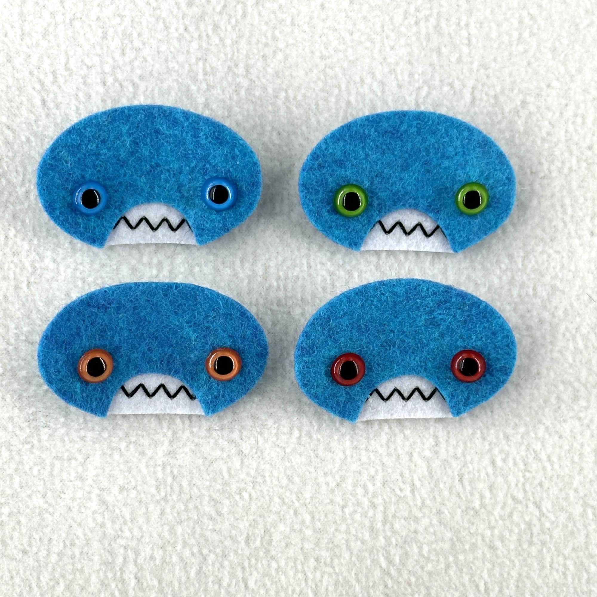 Four oval shaped teal faces with different eye colors and pointed teeth sit atop a white fleece background.